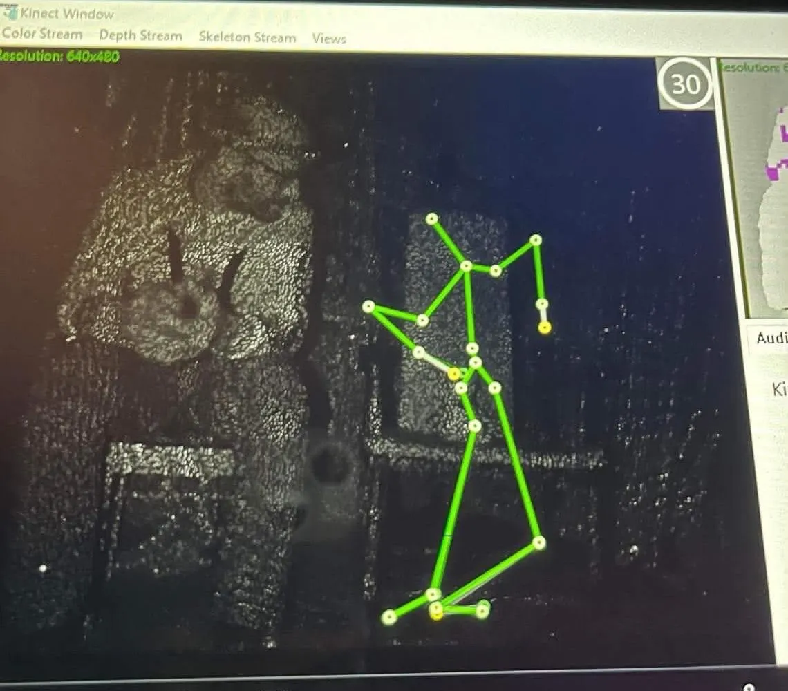 Skjermbilde fra en Kinect som tegner opp et grønt skjelett ved siden av en stol i et mørkt rom