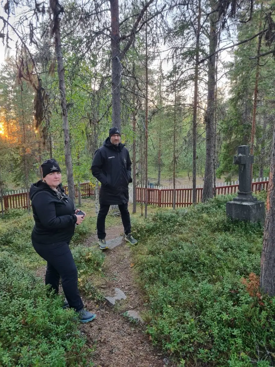 To personer i svarte klær står ved en inngjerdet skogskirkegård med et steinkors, i kveldssol