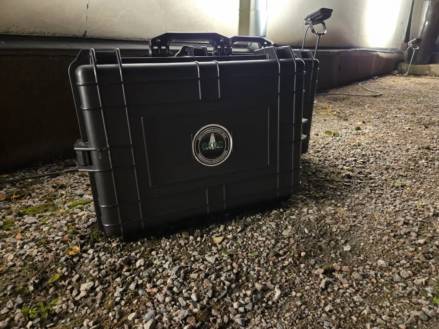 A black flight case bearing the GKGC emblem standing on gravel beneath a building