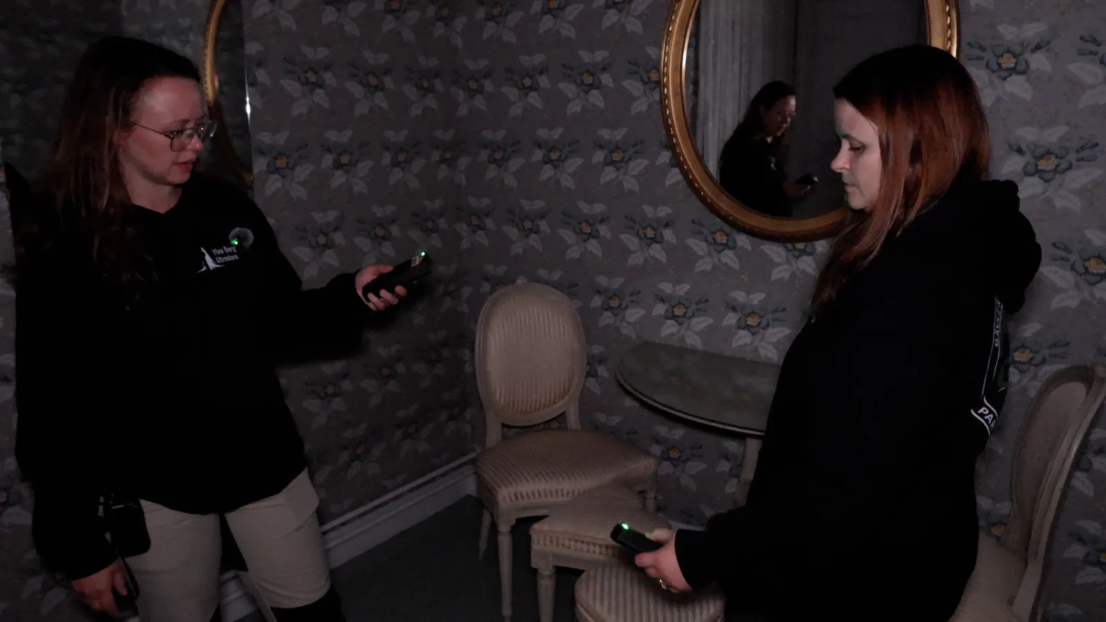 Linnéa and Ylva facing each other, each holding a meter, in a wallpapered room with a mirror
