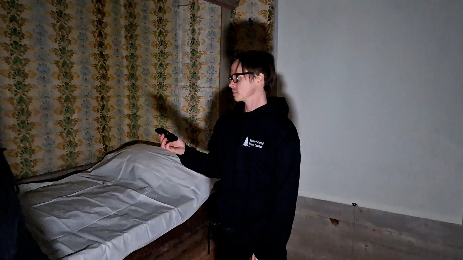 Robert holding out a meter towards an old bed in a floral-wallpapered room